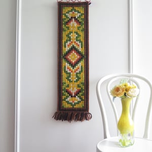 Può includere: Un arazzo rettangolare da parete con un disegno geometrico nei toni del giallo, verde, rosso e marrone. L'arazzo ha un bordo marrone e frange sul fondo. Un vaso giallo con fiori è su una sedia bianca.