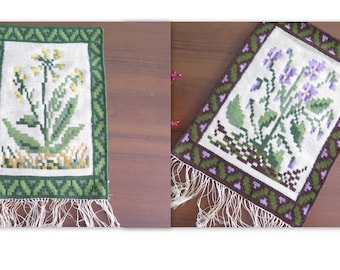 Puede incluir: Dos tapices de punto de aguja enmarcados con diseños florales. El primero presenta una flor amarilla con hojas verdes y un borde marrón y verde. El segundo presenta una flor morada con hojas verdes y un borde marrón, verde y morado.