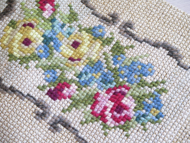 May include: Close-up of a beige fabric with embroidered flowers in shades of pink, red, yellow, blue, and green. The flowers are arranged in a symmetrical pattern.