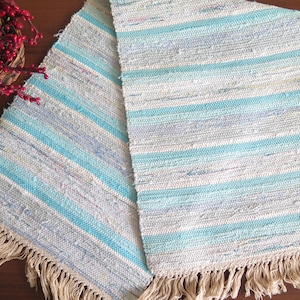 May include: A blue and white striped woven rug with fringe on the edges. The rug is made of natural fibres and has a rustic look.