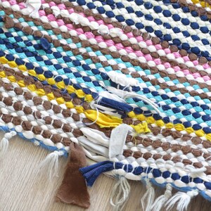 May include: A colorful woven rug with a frayed edge and loose threads. The rug is made of different colored fabrics, including brown, white, blue, yellow, and pink.