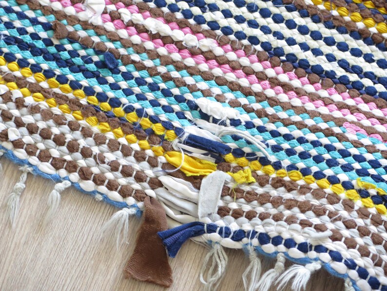 May include: A colorful woven rug with a frayed edge and loose threads. The rug is made of different colored fabrics, including brown, white, blue, yellow, and pink.