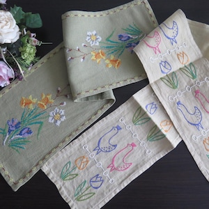 Könnte beinhalten: Drei bestickte Tischläufer. Ein Läufer zeigt ein Blumenmuster mit gelben, orangefarbenen und violetten Blumen. Die beiden anderen Läufer zeigen ein sich wiederholendes Muster aus Vögeln und Blumen in Rosa, Blau und Grün.