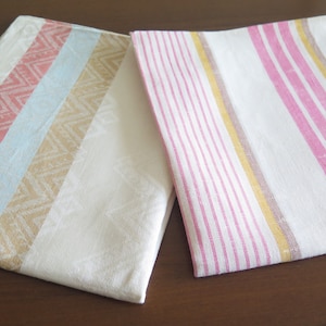May include: Two white kitchen towels with different patterns. One towel has pink, blue, and gold stripes. The other towel has pink, blue, and gold stripes with a chevron pattern.