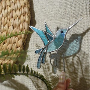 Puede incluir: Una escultura de colibrí de vidrieras en tonos azules y turquesas, con un largo pico plateado y un ojo negro. El pájaro está montado sobre un alambre de metal, y las alas están extendidas en vuelo. El fondo incluye una cesta tejida y follaje verde.