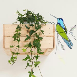 Puede incluir: Una jardinera de caja de madera con hiedra verde en cascada. Un colibrí de vidriera azul y verde se posa cerca. La jardinera está montada en una pared blanca, creando una exhibición decorativa natural.