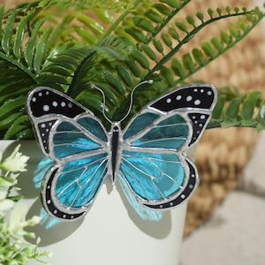 Puede incluir: Una mariposa de vidriera turquesa con detalles en blanco y negro y un contorno plateado. La mariposa está colocada sobre un fondo de follaje verde. Las alas están detalladas con líneas, para decoración.