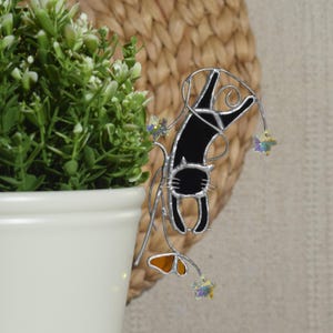 May include: A black cat-shaped garden stake with a silver metal frame. The cat is hanging from a vine with orange and crystal-like accents. The stake is next to a white potted plant with green foliage.