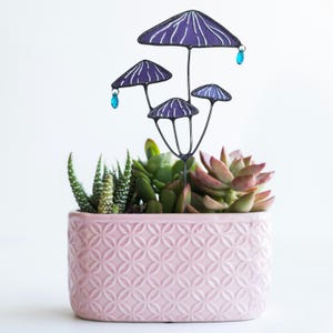 May include: A pink rectangular planter with a geometric pattern, filled with succulents. A metal mushroom stake with purple caps and blue crystal accents is placed in the planter, creating a whimsical garden accent.
