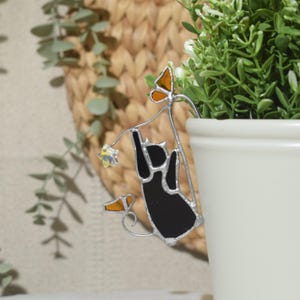 May include: A black cat-shaped plant stake with a silver outline, holding onto a star. The stake has orange and yellow butterfly accents. The cat is hanging from a stem, with a white pot and green plant in the background.