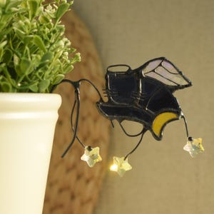 Könnte beinhalten: Glasmalerei-Fledermaus-Dekoration mit dunkelblauem Körper, gelben Akzenten und schillernden Flügeln. Die Fledermaus hängt an einem schwarzen Draht mit drei sternförmigen Kristallen. Die Dekoration befindet sich in der Nähe eines weißen Blumentopfs mit grünen Blättern.