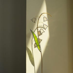 Op de afbeelding: Een sculptuur van glas-in-lood in de vorm van een lelietje-van-dalen met een lange, dunne messing steel, een enkel, gestructureerd, felgroen blad en verschillende kleine, doorschijnende, witte klokvormige bloemen met donkere metalen randen.