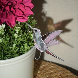 May include: A stained glass hummingbird with pink, brown, and silver details perches near a pink flower and green foliage. The bird has outstretched wings and a long beak. The hummingbird is attached to a metal wire.