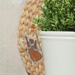 May include: A stained glass cat decoration in shades of brown and orange, reaching for a star. The cat is attached to a plant pot with a white ceramic finish. The background features a woven texture and greenery.