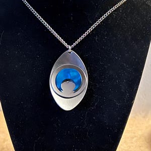 May include: A silver chain necklace with a pendant featuring a blue crescent moon design. The pendant is oval shaped and has a brushed metal finish.