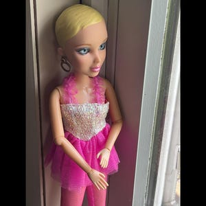 May include: A fashion doll with blonde hair and blue eyes, wearing a pink dress with a sequined bodice and matching tights. The doll has hoop earrings and is posed near a window.