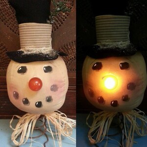 May include: A snowman figurine with a black top hat, a red nose, and black eyes. The snowman is sitting on a spring and has a straw skirt. The snowman's nose is lit up, creating a warm glow.
