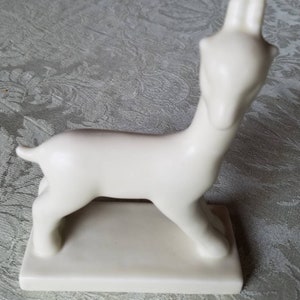 Könnte beinhalten: Eine weiße Keramikfigur eines stilisierten Hirsches mit langem Hals und schlanken Beinen. Der Hirsch steht auf einem rechteckigen Sockel.