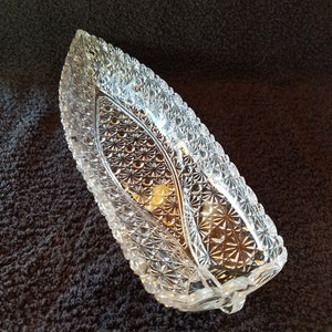 Op de afbeelding: Een heldere glazen serveerschaal met een decoratief, geslepen glaspatroon. De schaal is gevormd als een lang, smal bootje.