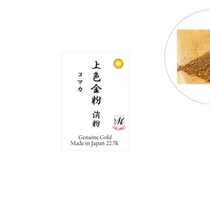 Op de afbeelding: Een klein wit label met Japanse tekst die "上金粉" (wat "gouden poeder" betekent) en "コマカ" (wat "fijn" betekent) leest. Het label vermeldt ook "Genuine Gold Made in Japan 22.7k". Op de achtergrond is een kleine hoop gouden poeder te zien.