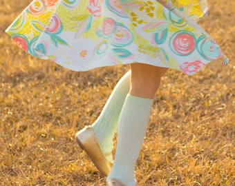 Puede incluir: Una persona joven que lleva una falda floral colorida y calcetines hasta la rodilla de color verde claro con zapatos planos dorados. La persona está de pie en un campo de hierba seca.