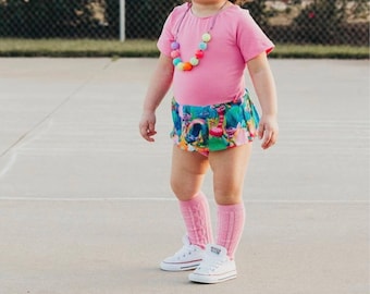 Puede incluir: Un niño pequeño vestido de troll para Halloween. Lleva una camisa rosa, una falda colorida con estampado de troll, calcetines rosas hasta la rodilla y zapatillas blancas. Lleva una peluca rosa de troll con orejas en la cabeza y un collar de cuentas de colores.
