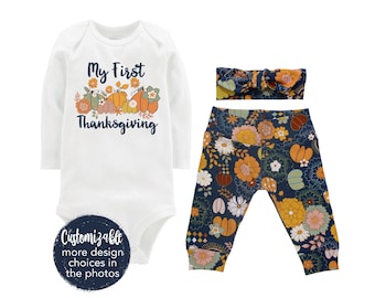 May include: A white long-sleeved bodysuit with the text "My First Thanksgiving" and a graphic of pumpkins and fall leaves. A pair of blue leggings with a floral pattern and a blue headband with a floral pattern.