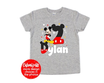 Puede incluir: Camiseta gris de manga corta con un personaje de Mickey Mouse rojo y negro que lleva una camisa roja y zapatos amarillos. El número 2 está detrás de Mickey Mouse y el nombre Dylan está escrito debajo. La camiseta tiene un diseño de estrella blanca y confeti amarillo.