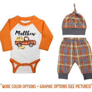 May include: A baby outfit set with a white bodysuit featuring a red truck with pumpkins, the name "Matthew" and orange sleeves, plaid pants, and a plaid hat.