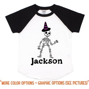 May include: White and black baseball tee shirt with a skeleton wearing a purple and black witch hat. The text "Jackson" is printed below the skeleton.