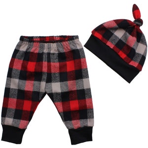 Op de afbeelding: Rode, zwarte en grijze geruite babybroek en muts set. De broek heeft een elastische tailleband en gezoomde pijpen. De muts heeft een geknoopte bovenkant.