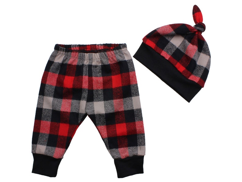 Op de afbeelding: Rode, zwarte en grijze geruite babybroek en muts set. De broek heeft een elastische tailleband en gezoomde pijpen. De muts heeft een geknoopte bovenkant.