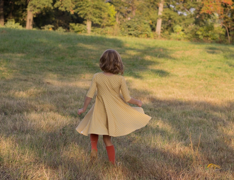May include: A young person wearing a yellow dress with white polka dots and red tights spins in a grassy field.