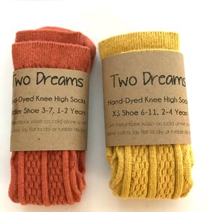 May include: Two pairs of hand-dyed knee-high socks, one pair is orange and the other is yellow. The socks are folded and have a brown paper tag with the words "Two Dreams" and care instructions.