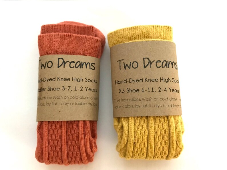 May include: Two pairs of hand-dyed knee-high socks, one pair is orange and the other is yellow. The socks are folded and have a brown paper tag with the words "Two Dreams" and care instructions.