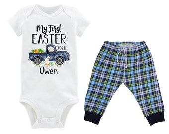 Puede incluir: Un body blanco para bebé con el texto "My First Easter 2020" y un camión azul con un conejito. El body se combina con pantalones de cuadros azules y verdes.