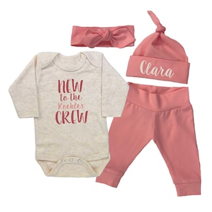 Op de afbeelding: Een roze en witte baby outfit set met een longsleeve bodysuit met de tekst "New to the Koehler Crew", een roze hoofdband, een roze muts met de naam "Clara" erop geborduurd en roze leggings.