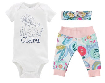 Könnte beinhalten: Ein weißer Baby-Strampler mit einem Hasen- und Blumendesign und dem Namen "Clara" in Blau bedruckt. Der Strampler wird mit pinkfarbenen und weißen Leggings mit Blumenmuster und einem pinkfarbenen und weißen Blumen-Stirnband kombiniert.