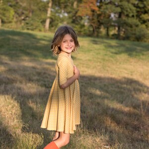 May include: A young girl wearing a yellow polka dot dress and red knee-high socks stands in a grassy field. She is smiling and looking at the camera.