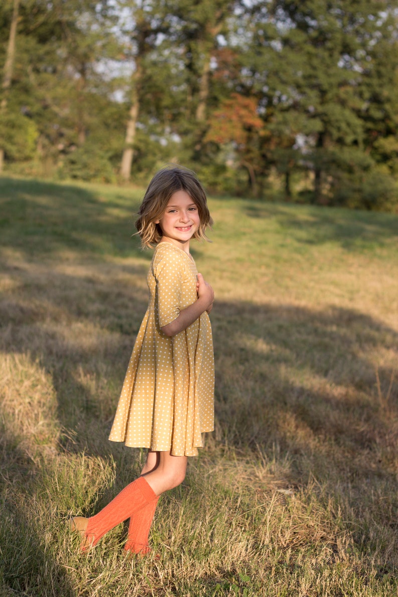 May include: A young girl wearing a yellow polka dot dress and red knee-high socks stands in a grassy field. She is smiling and looking at the camera.