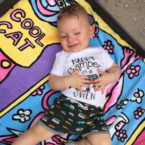 May include: A baby boy wearing a white onesie with the text "Happy Camper Owen" and a black and white patterned shorts with a camper van design. The baby is lying on a colorful beach towel with the text "Cool Cat" on it.