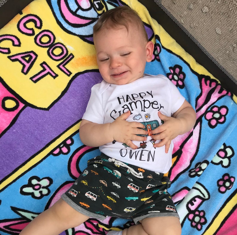 May include: A baby boy wearing a white onesie with the text "Happy Camper Owen" and a black and white patterned shorts with a camper van design. The baby is lying on a colorful beach towel with the text "Cool Cat" on it.