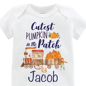 Może przedstawiać: Biały śpioszek dla niemowlęcia z niebiesko-pomarańczową grafiką pociągu przewożącego dynie. Tekst "Cutest Pumpkin in the Patch" znajduje się nad pociągiem, a imię "Jacob" pod pociągiem.