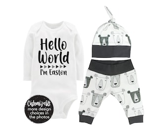 Könnte beinhalten: Ein weißer Baby-Strampler mit langen Ärmeln und dem Text "Hello World I'm Easton". Eine weiße und graue Mütze mit schwarzem Besatz und eine schwarze und weiße gemusterte Hose mit schwarzem Bund.