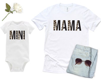 May include: White t-shirt with the word "MAMA" in black letters with a leopard print pattern. A white onesie with the word "MINI" in black letters with a leopard print pattern.