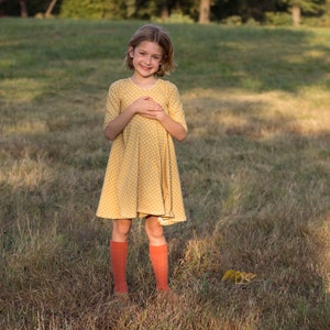May include: A young girl wearing a yellow polka dot dress and orange knee-high socks stands in a field of tall grass.