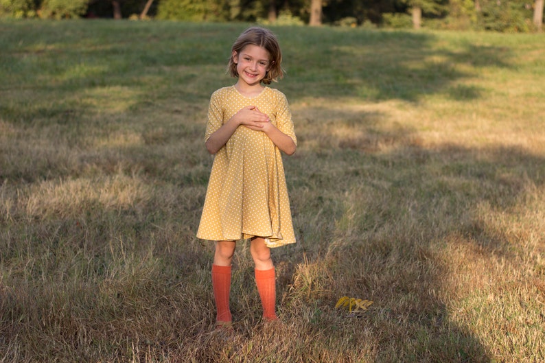 May include: A young girl wearing a yellow polka dot dress and orange knee-high socks stands in a field of tall grass.