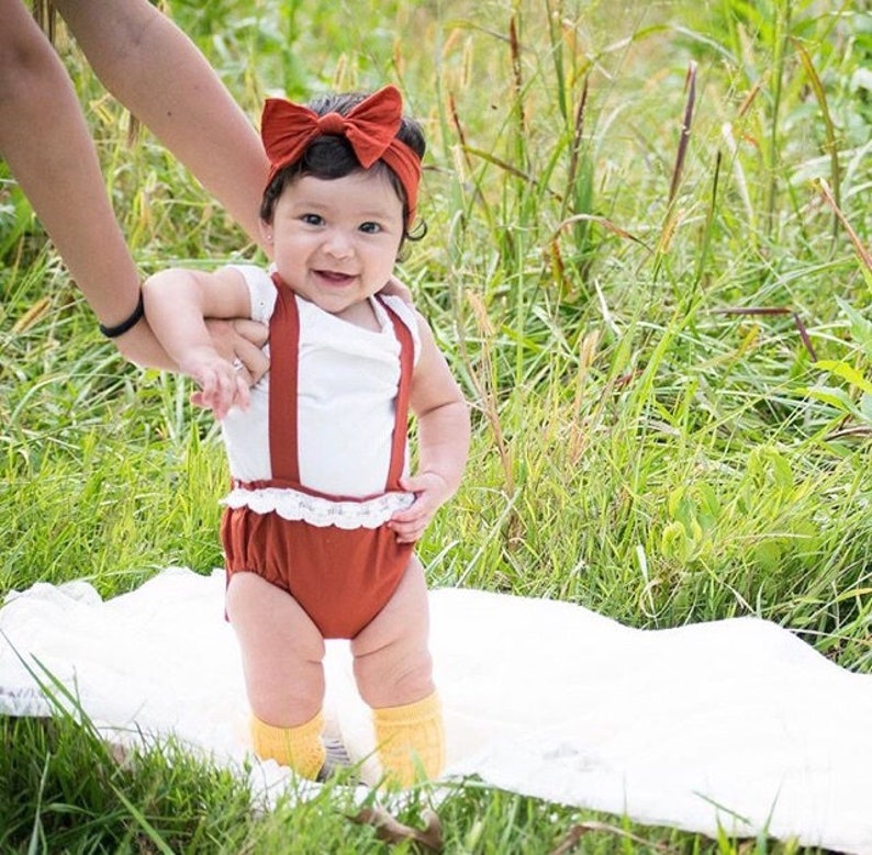 May include: A baby girl wearing a white sleeveless shirt with brown suspenders and a matching brown skirt with a white lace trim. She is wearing a brown headband with a bow and yellow socks. She is standing on a white blanket in a grassy field.
