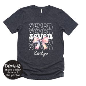 May include: A dark blue t-shirt with the word "seven" repeated in a retro font with a pink bow. The name "Evelyn" is printed below the design.