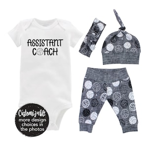 May include: A baby outfit set with a white bodysuit that says "Assistant Coach" with a volleyball graphic, a gray headband with a volleyball graphic, a gray hat with a volleyball graphic, and gray leggings with a volleyball graphic.